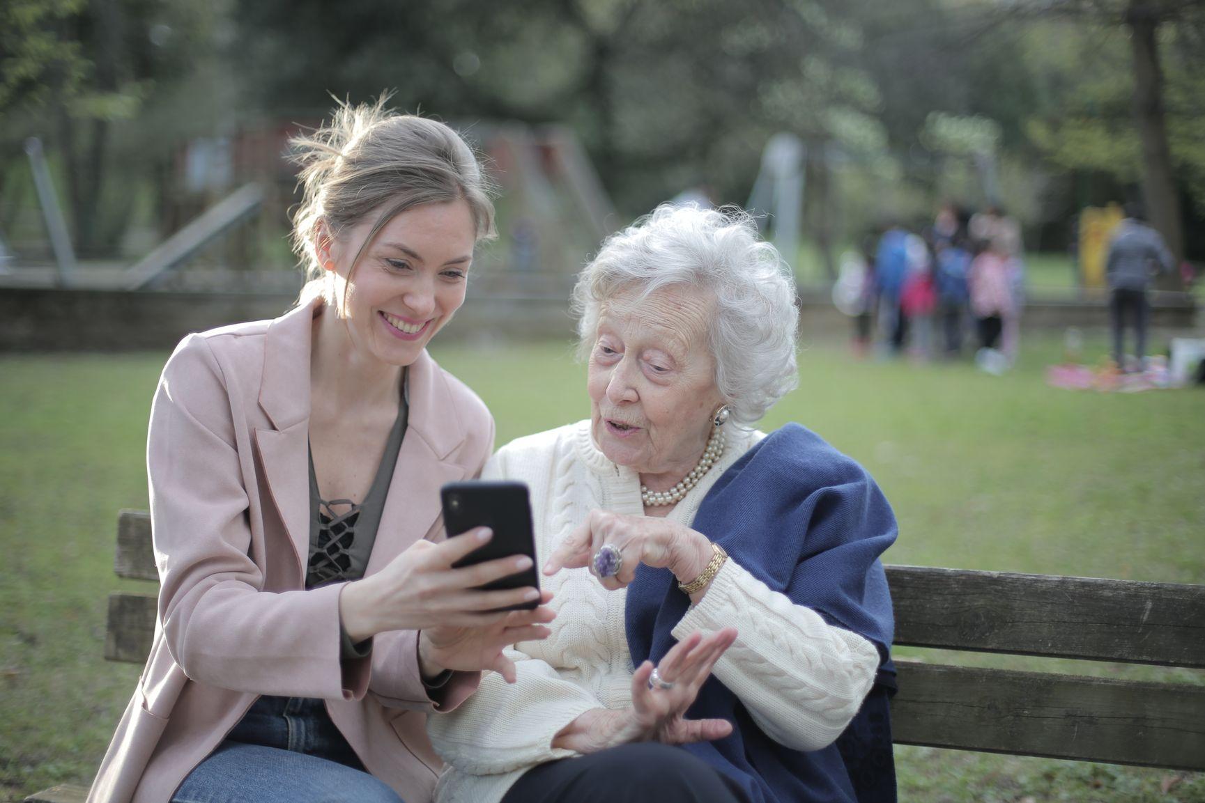 An adult daughter and her senior mother sitting on a park bench together, looking closely at a smartphone screen